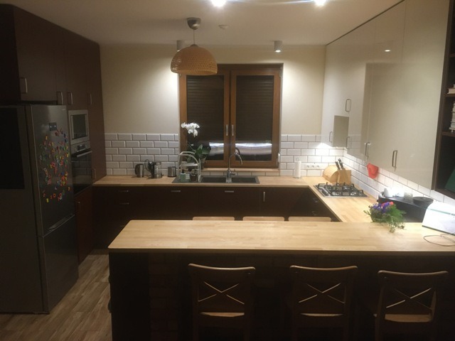 Nowoczesna kuchnia z drewnianym blatem, białymi płytkami metra i ciemnymi frontami szafek, oświetlona lampą wiszącą z wikliny.