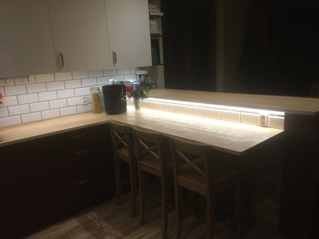 Kuchenny blat z drewnianym wykończeniem, oświetleniem LED podszafkowym i trzema drewnianymi krzesłami barowymi, widok na białe płytki metra i ciemne szafki.