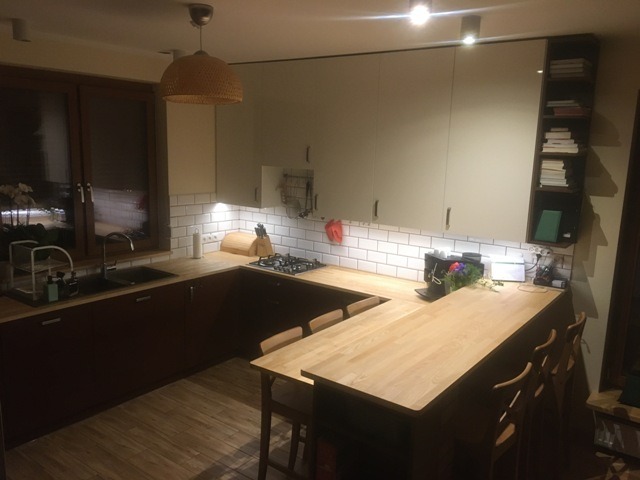 Ukształtowana w literę U kuchnia z drewnianym blatem i niskim barkiem, ciemnobrązowe dolne szafki i jasne górne, białe kafelki na ścianie, oświetlenie punktowe pod szafkami, widok na nocne miasto...