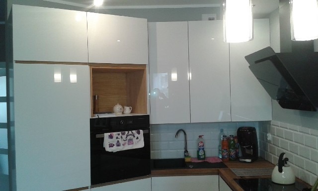 Nowoczesna kuchnia z białymi, lakierowanymi frontami szafek, czarnym piekarnikiem z ozdobną ściereczką, drewnianymi akcentami w otwartej półce i blacie, oraz czarnym okapem nad płytą grzewczą.