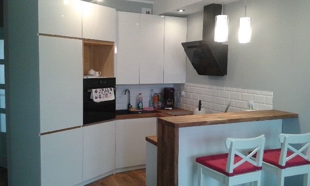 Nowoczesna kuchnia z białymi, błyszczącymi frontami szafek, drewnianymi akcentami, czarnym piekarnikiem i okapem oraz barkiem z białymi krzesłami barowymi z czerwonymi siedziskami.