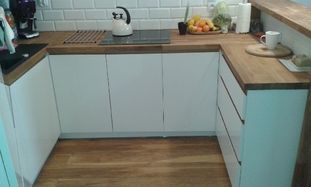 U-kształtna zabudowa kuchenna z białymi, gładkimi frontami i drewnianym blatem, na którym stoją czajnik, owoce i kubek.