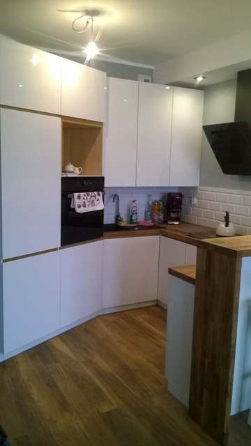 Nowoczesna kuchnia z białymi, lakierowanymi frontami szafek, drewnianym blatem i parkietem, z czarnym piekarnikiem i okapem oraz akcesoriami kuchennymi na blacie.