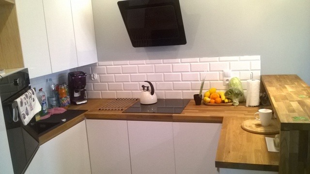 Nowoczesna kuchnia z białymi frontami, drewnianym blatem, białymi płytkami metra i czarnym okapem, na blacie czajnik i owoce.