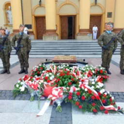 Wieniec z czerwono-białych kwiatów u stóp pomnika, strzeżony przez czterech żołnierzy w mundurach na tle klasycystycznego budynku z kolumnami.