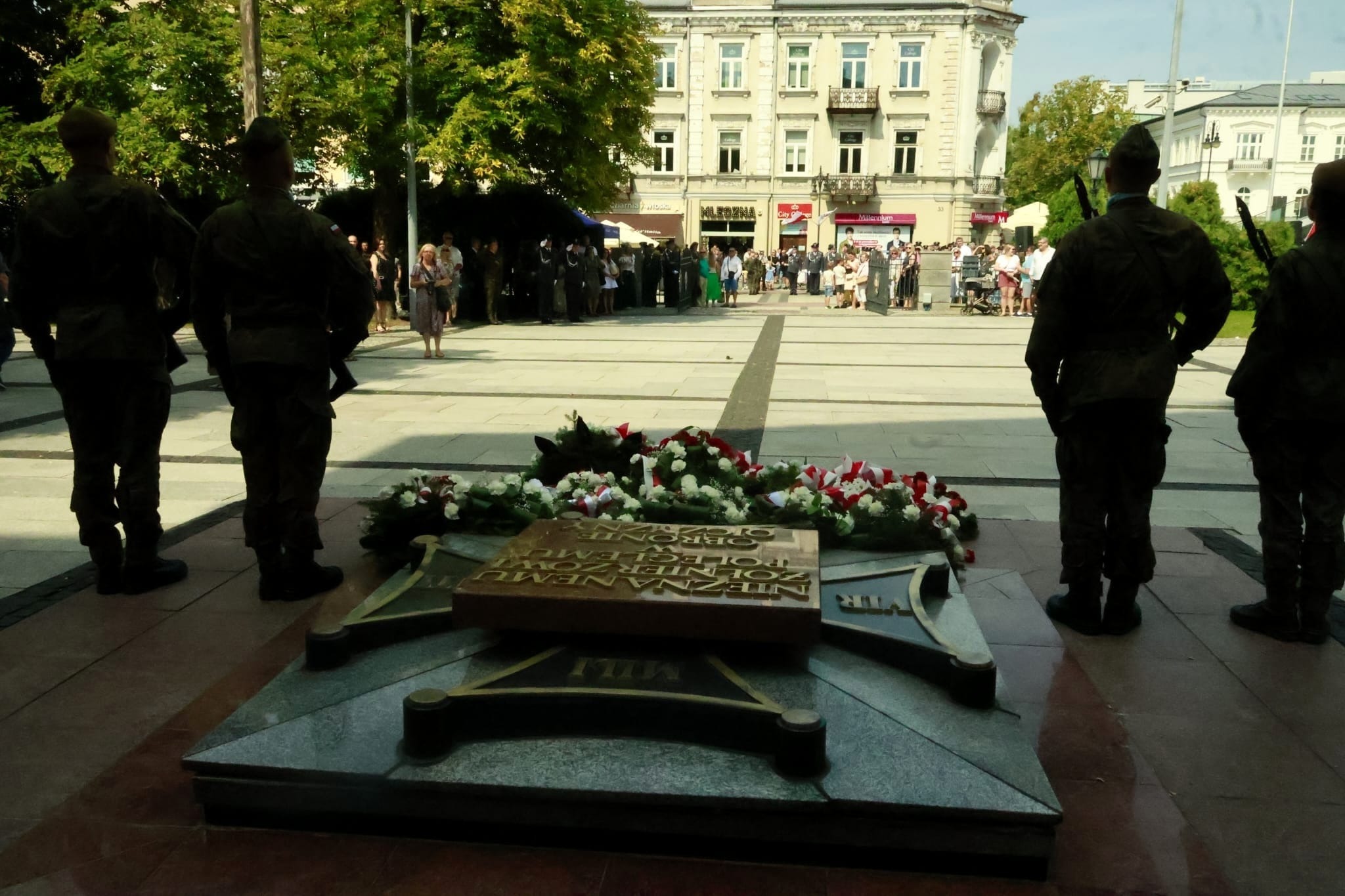 Żołnierze w mundurach stojący na warcie przy Grobie Nieznanego Żołnierza, otoczonym wieńcami z biało-czerwonymi kwiatami, w tle zgromadzenie ludzi na placu miejskim.