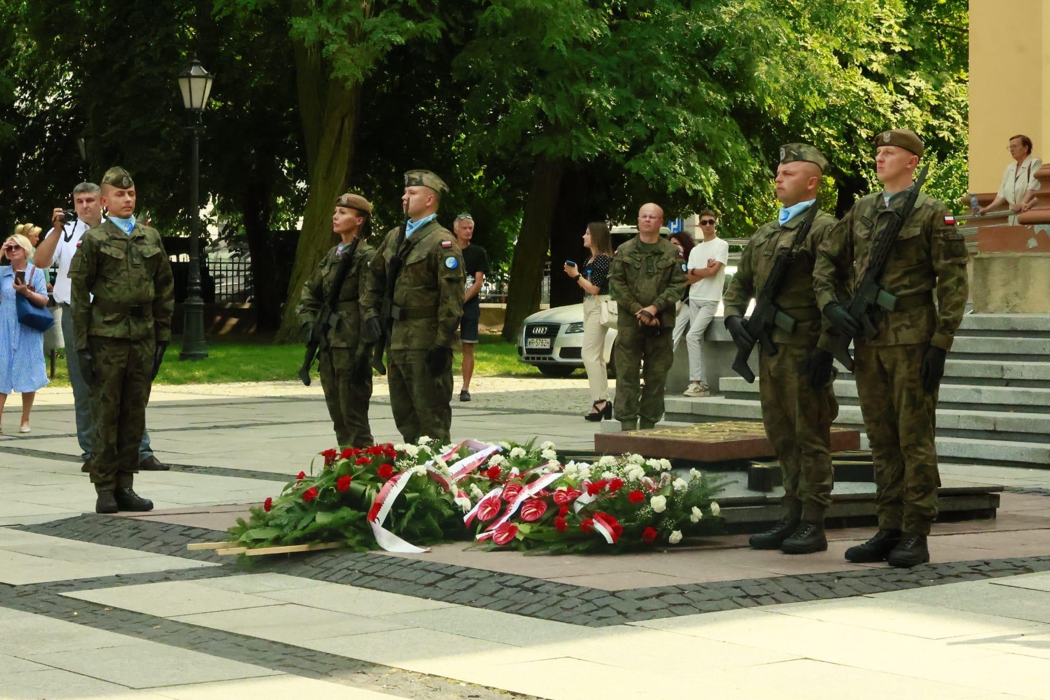 Żołnierze w mundurach galowych stojący na placu przed pomnikiem z wieńcami w barwach narodowych.