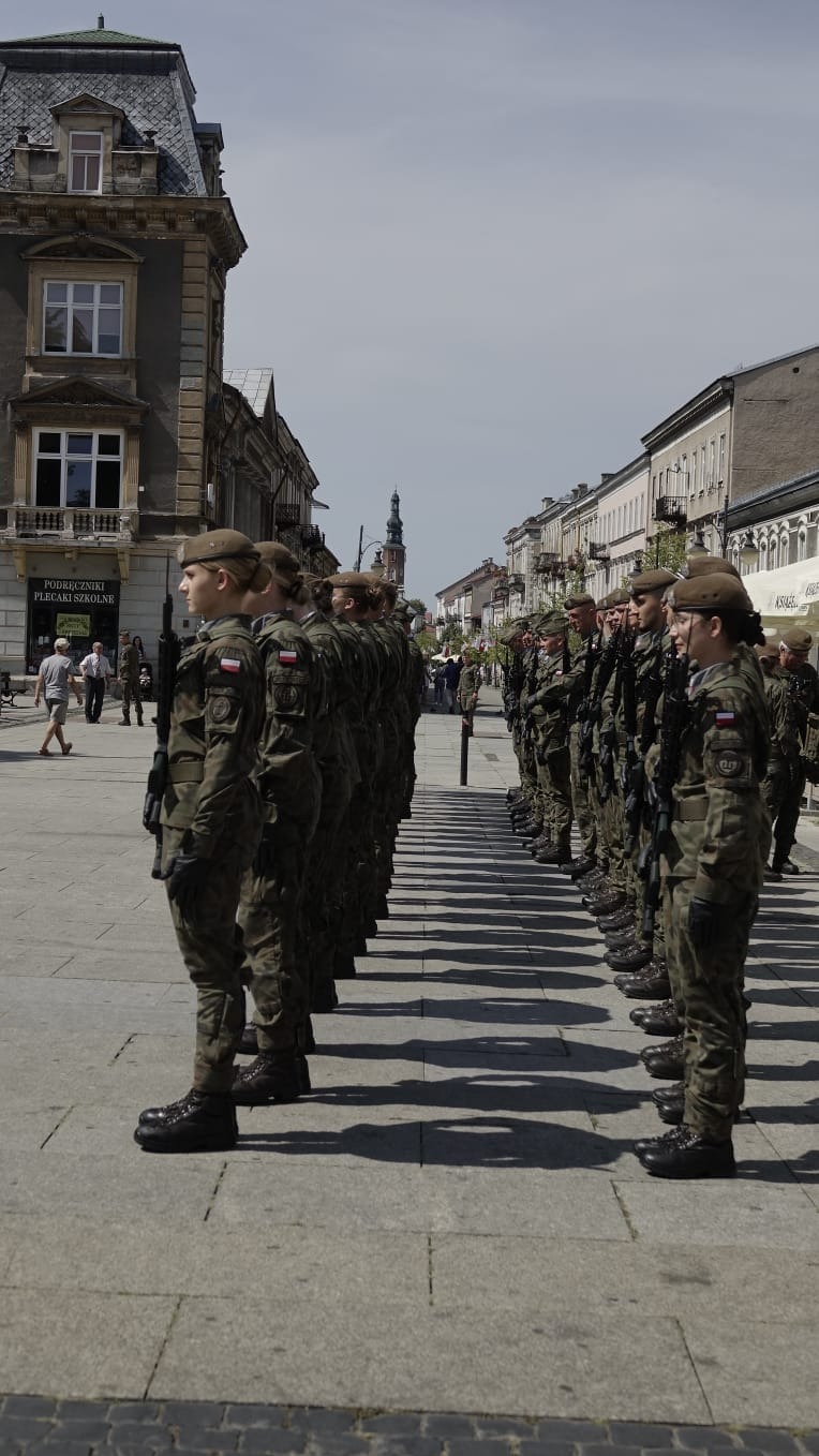 Długi szereg umundurowanych żołnierzy Wojska Polskiego w beretach, stojących na placu w mieście, z flagami Polski na ramionach, w tle kamienice i wieża kościoła.