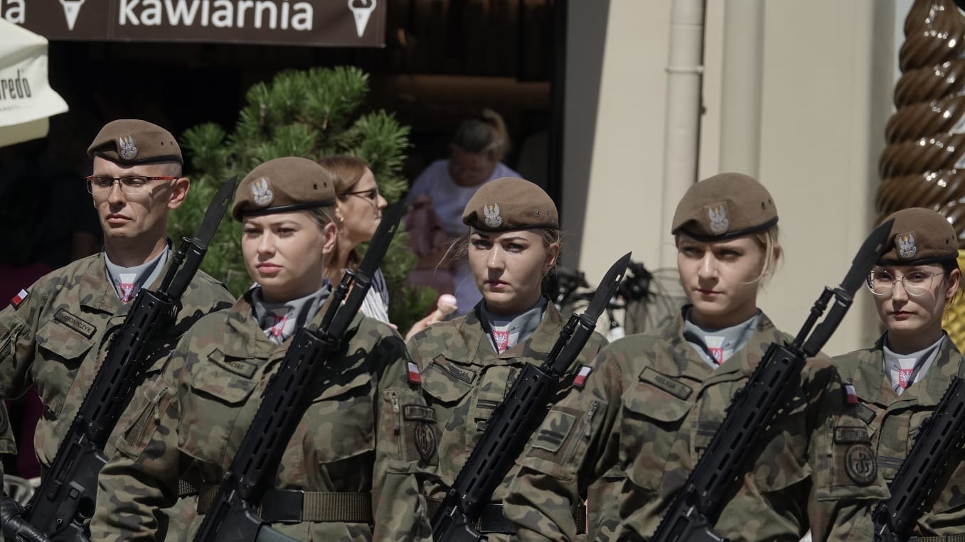 Grupa żołnierzy Wojska Polskiego w umundurowaniu polowym, z karabinami, podczas uroczystości plenerowej, w tle widoczna kawiarnia.