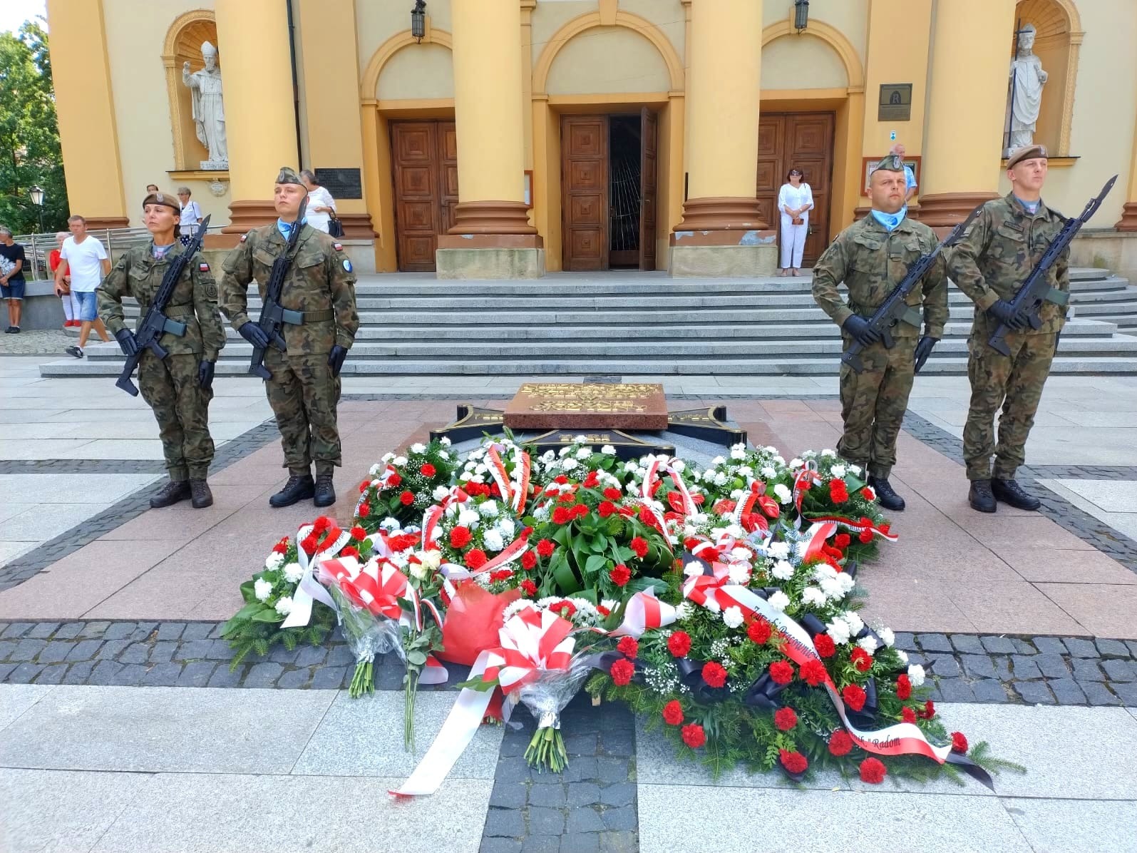 Wieniec z czerwono-białych kwiatów u stóp pomnika, strzeżony przez czterech żołnierzy w mundurach na tle klasycystycznego budynku z kolumnami.
