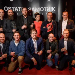 Grupowe zdjęcie ekipy filmowej z szampanem na tle plakatu 'Ostatni Samotnik' z widocznymi logotypami partnerów.