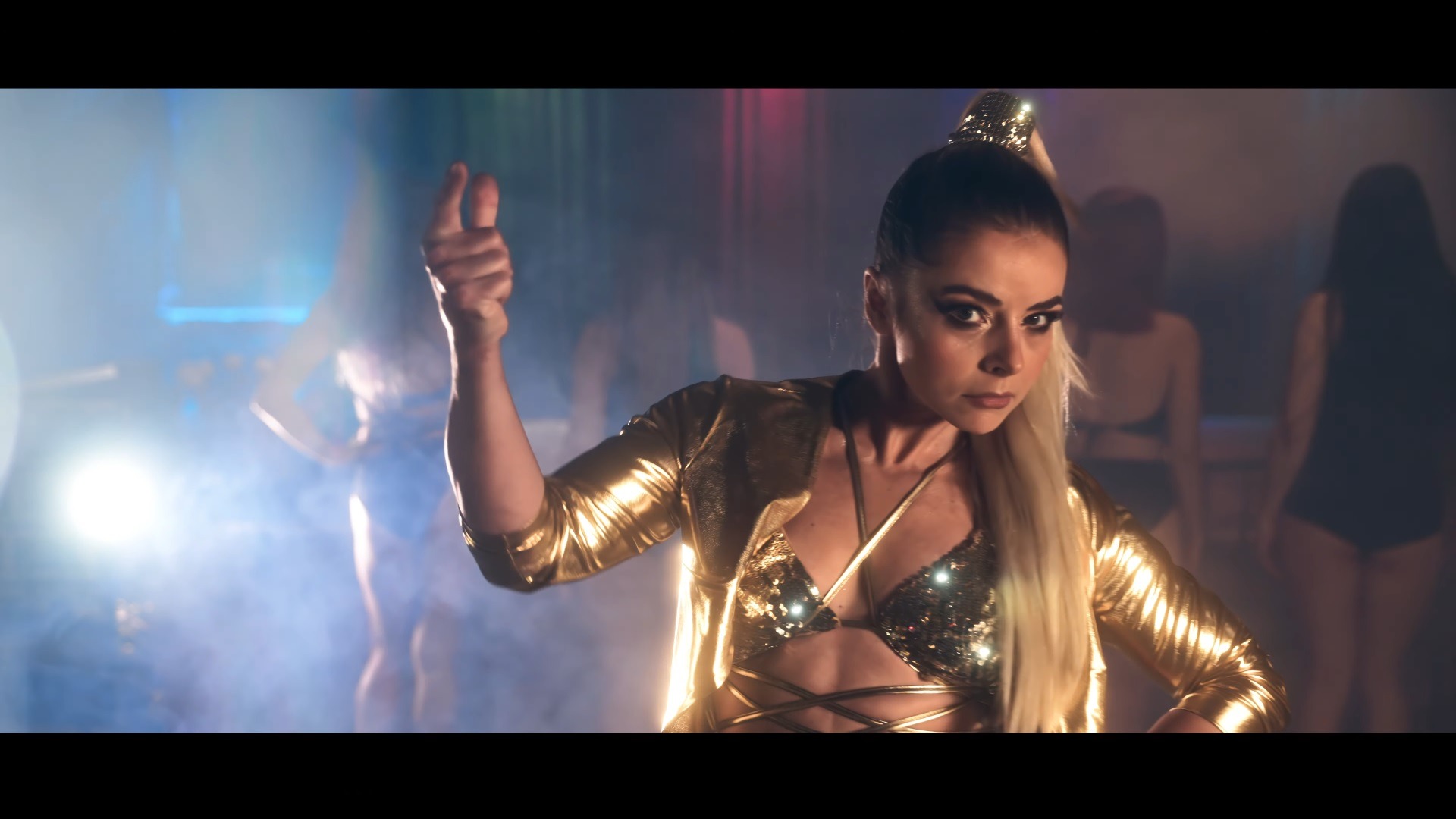 Kobieta w złotej kurtce i cekinowym topie, z blond włosami upiętymi w wysoki kucyk, w pozie tanecznej na tle innych tancerek i efektów świetlnych.