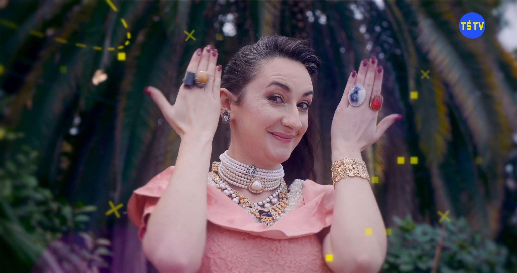 Kobieta w różowej sukience i bogatej biżuterii, z rękami uniesionymi do góry, prezentująca pierścionki na tle zieleni, z dodatkowymi efektami graficznymi w tle i logo TSTV w prawym górnym rogu.