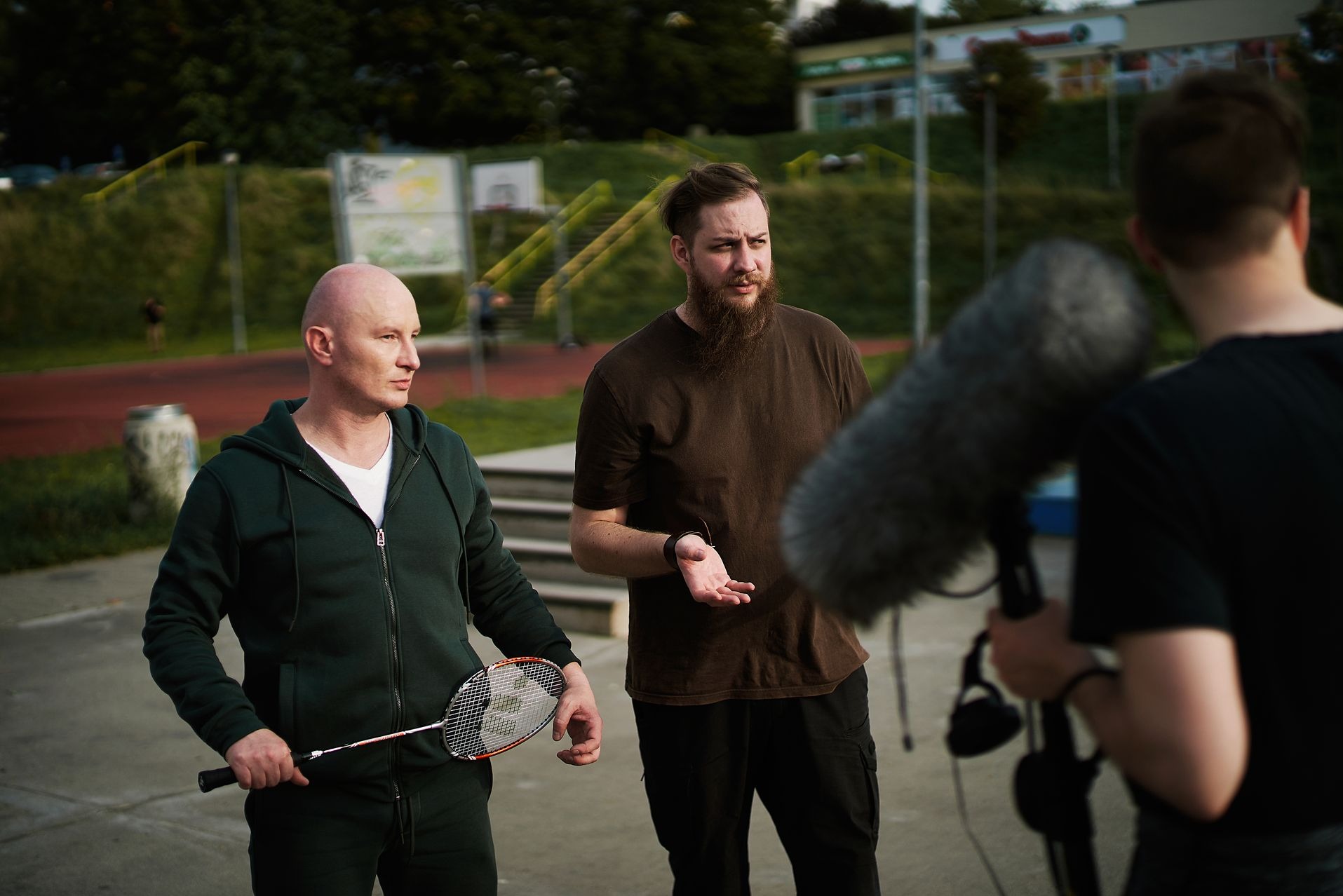 Ujęcie z planu filmowego: mężczyzna z rakietką do badmintona i mężczyzna z brodą rozmawiają z osobą trzymającą mikrofon typu 'dead cat', w tle boisko sportowe.