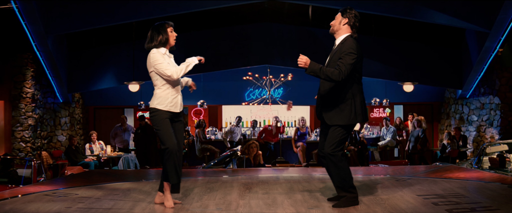 Scena taneczna z filmu, mężczyzna w garniturze i kobieta w białej koszuli, na tle baru z neonami i publicznością.