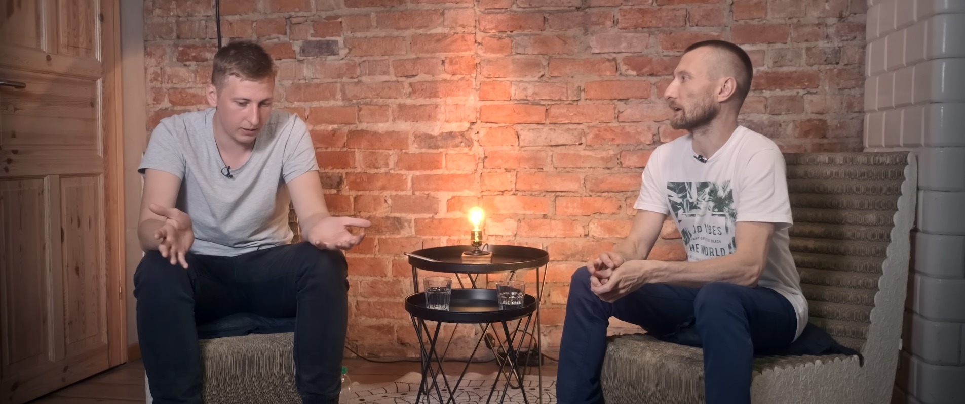 Dwóch mężczyzn siedzi w studio z ceglaną ścianą w tle, prowadząc rozmowę przy stoliku z zapaloną lampą i szklankami wody.