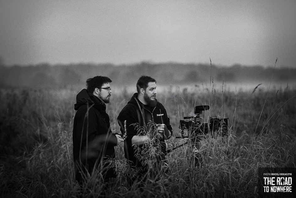 Czarno-białe ujęcie z planu filmowego w plenerze; dwóch mężczyzn stoi w wysokiej trawie obok kamery na statywie, w tle zarys lasu i pochmurne niebo.