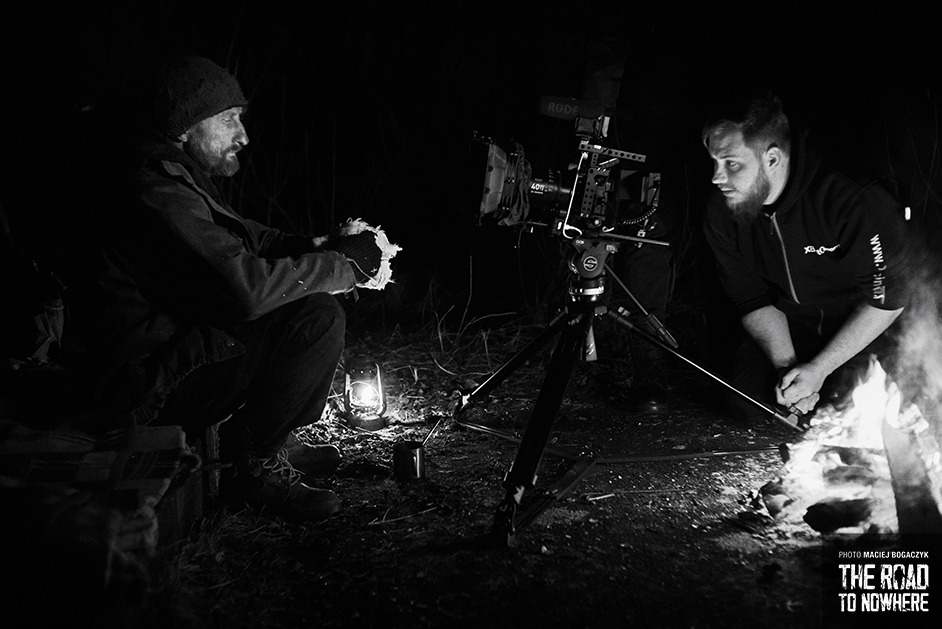 Czarno-białe zdjęcie z planu filmowego w nocy: dwóch mężczyzn przy ognisku, kamera na statywie z mikrofonem Rode, obok lampa naftowa, w tle las, na dole kadru widoczne logo filmu 'The Road to...