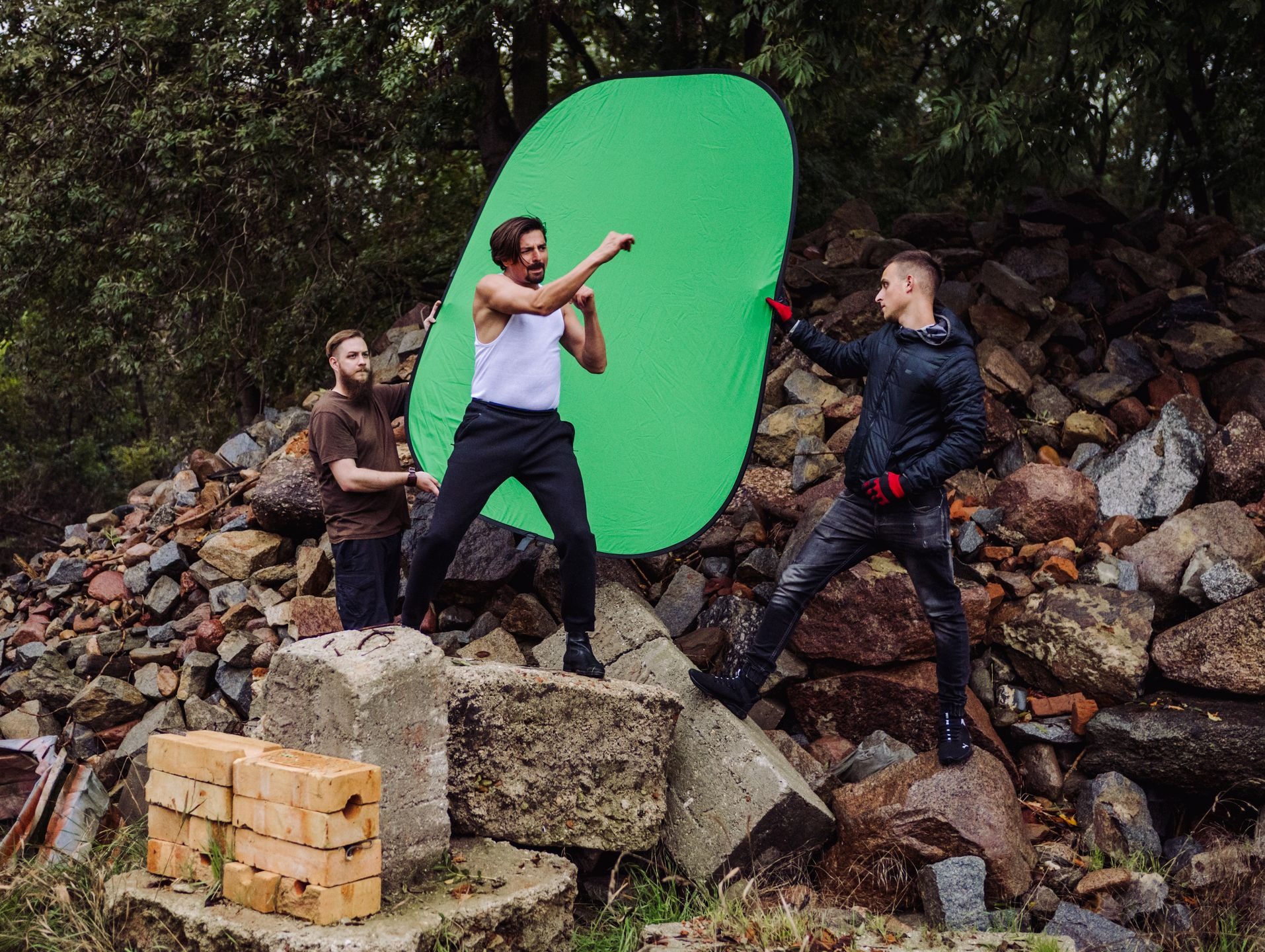 Zdjęcie zza kulis kręcenia materiału dla kabaretu Paranienormalni i Polskiej Nocy Kabaretowej: aktor w białym podkoszulku w pozie bokserskiej na tle zielonego ekranu, trzymanego przez dwie osoby...