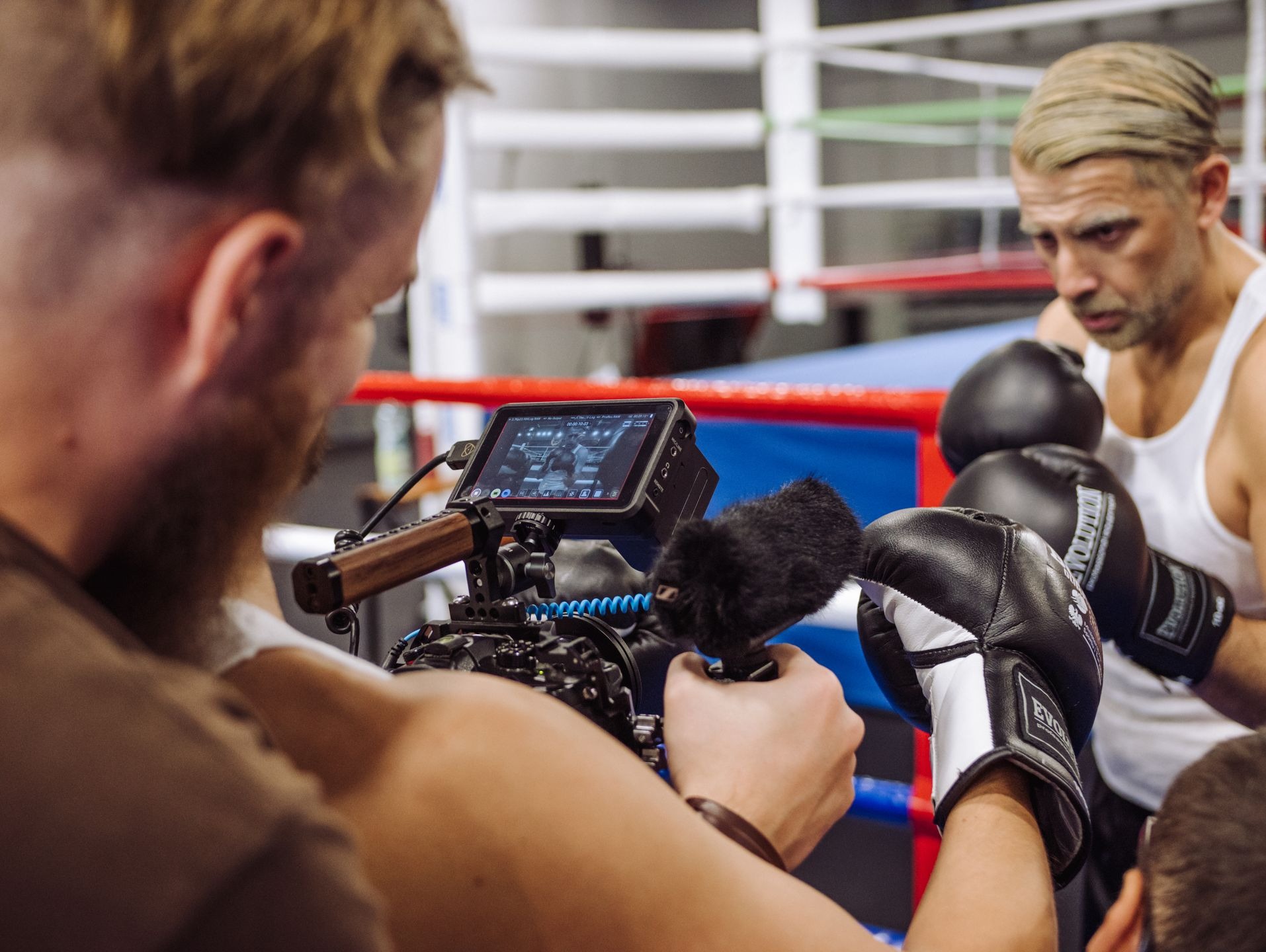 Ekipa filmowa kręci ujęcia boksera na ringu, widoczny monitor kamery z podglądem nagrywanego materiału i mikrofon na tyczce.