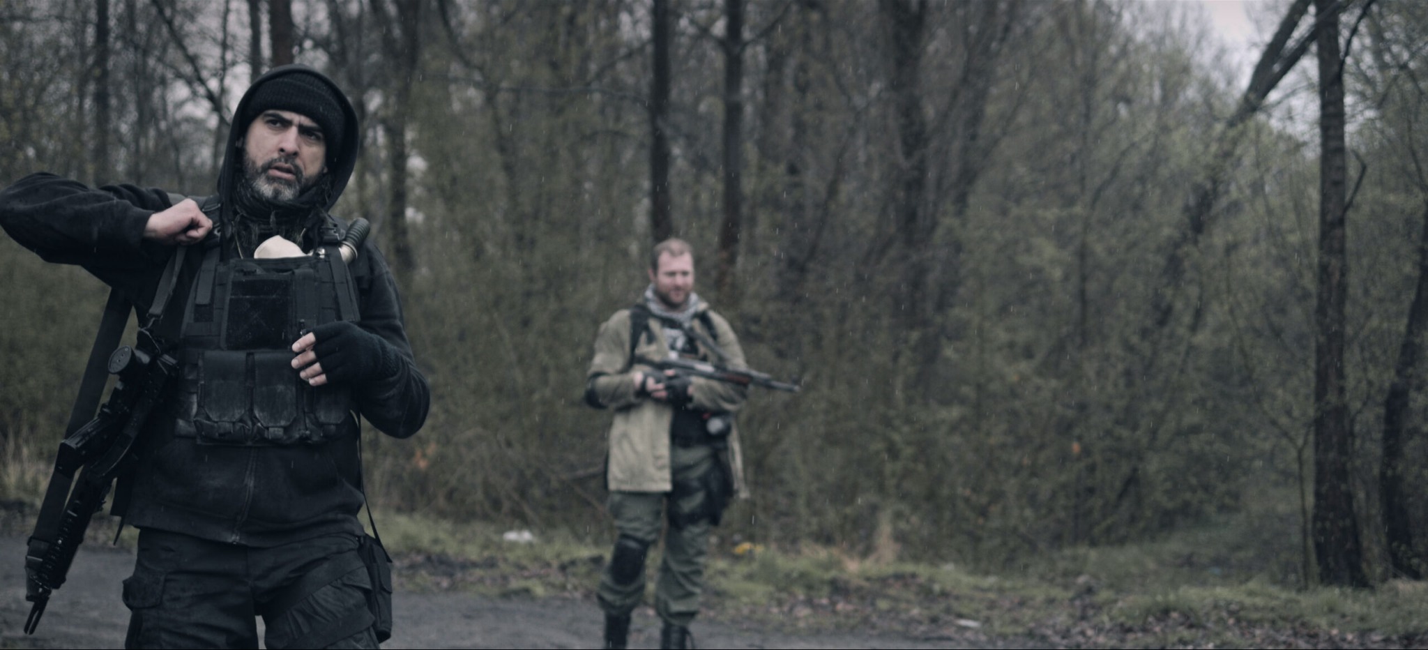 Scena z filmu fabularnego: Dwóch mężczyzn w taktycznym ubraniu i z bronią w zalesionym terenie podczas deszczu. Jeden z nich zapina kamizelkę taktyczną, drugi stoi w tle z karabinem.