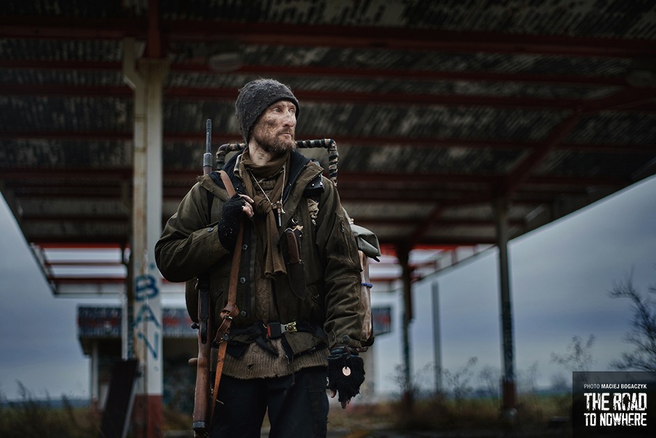 Mężczyzna z brodą i karabinem, ubrany w postapokaliptyczny strój, stoi na tle opuszczonej stacji benzynowej z graffiti, kadrowanie od dołu w pochmurny dzień.