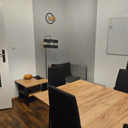 KANCELARIA PODATKOWA RENATA SMOLEŃ SPÓŁKA Z OGRANICZONĄ ODPOWIEDZIALNOŚCIĄ - Jasne biuro z drewnianym stołem, czarnymi krzesłami, lampą podłogową i tablicą suchościeralną na ścianie. Na małym stoliku miseczka i czarne pudełko.