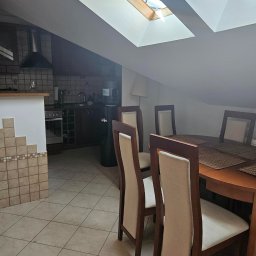 KANCELARIA PODATKOWA RENATA SMOLEŃ SPÓŁKA Z OGRANICZONĄ ODPOWIEDZIALNOŚCIĄ - Umeblowana jadalnia z drewnianym stołem, krzesłami z beżową tapicerką i widokiem na aneks kuchenny pod skośnym dachem z oknami dachowymi.