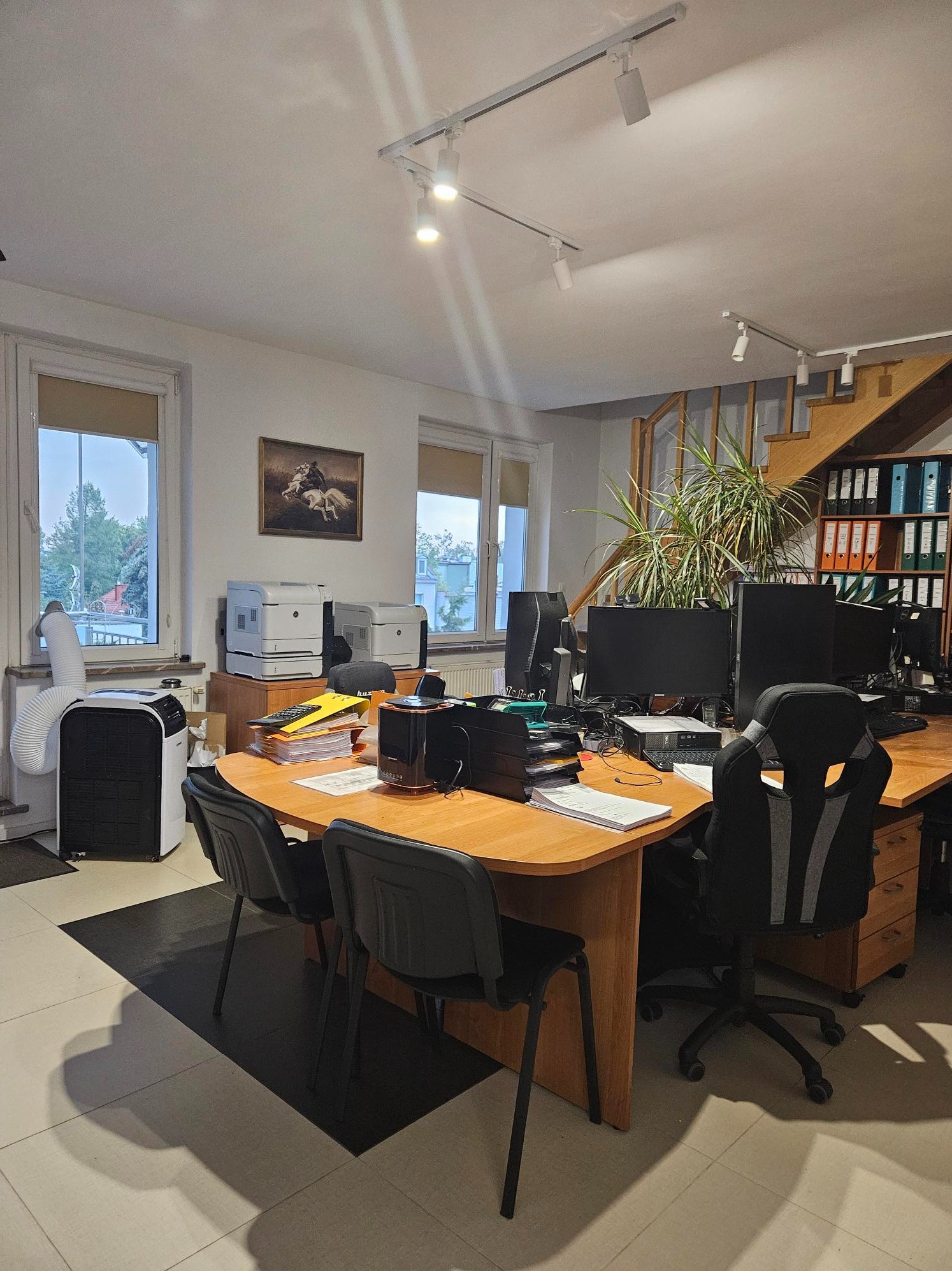 Jasne biuro z drewnianym biurkiem, komputerami, drukarkami, klimatyzatorem przenośnym i segregatorami na regale w tle. Na biurku leżą dokumenty i kalkulator.