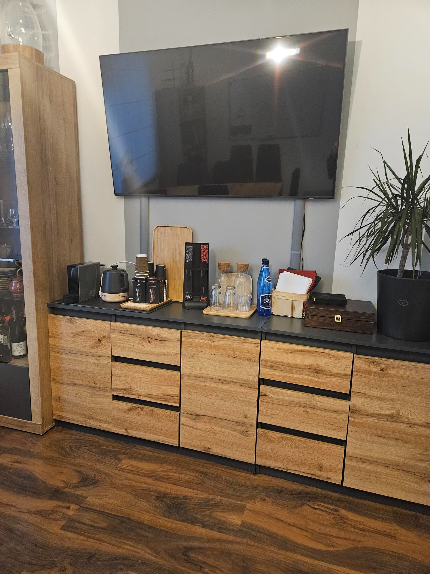 Nowoczesna komoda z drewnianymi frontami szuflad, na blacie ekspres do kawy, czajnik, szklanki, woda, dokumenty i roślina doniczkowa, nad nią telewizor.