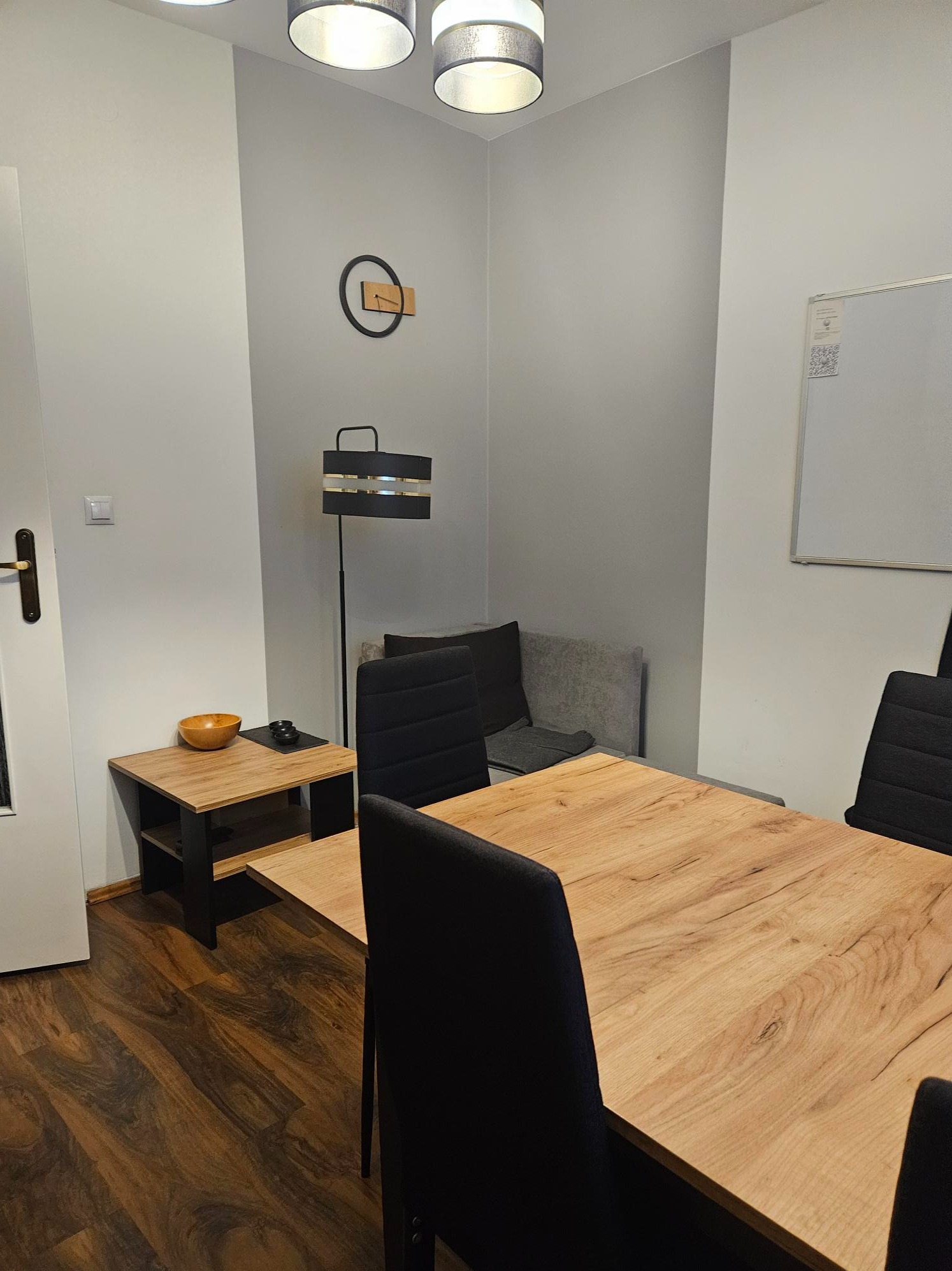Jasne biuro z drewnianym stołem, czarnymi krzesłami, lampą podłogową i tablicą suchościeralną na ścianie. Na małym stoliku miseczka i czarne pudełko.