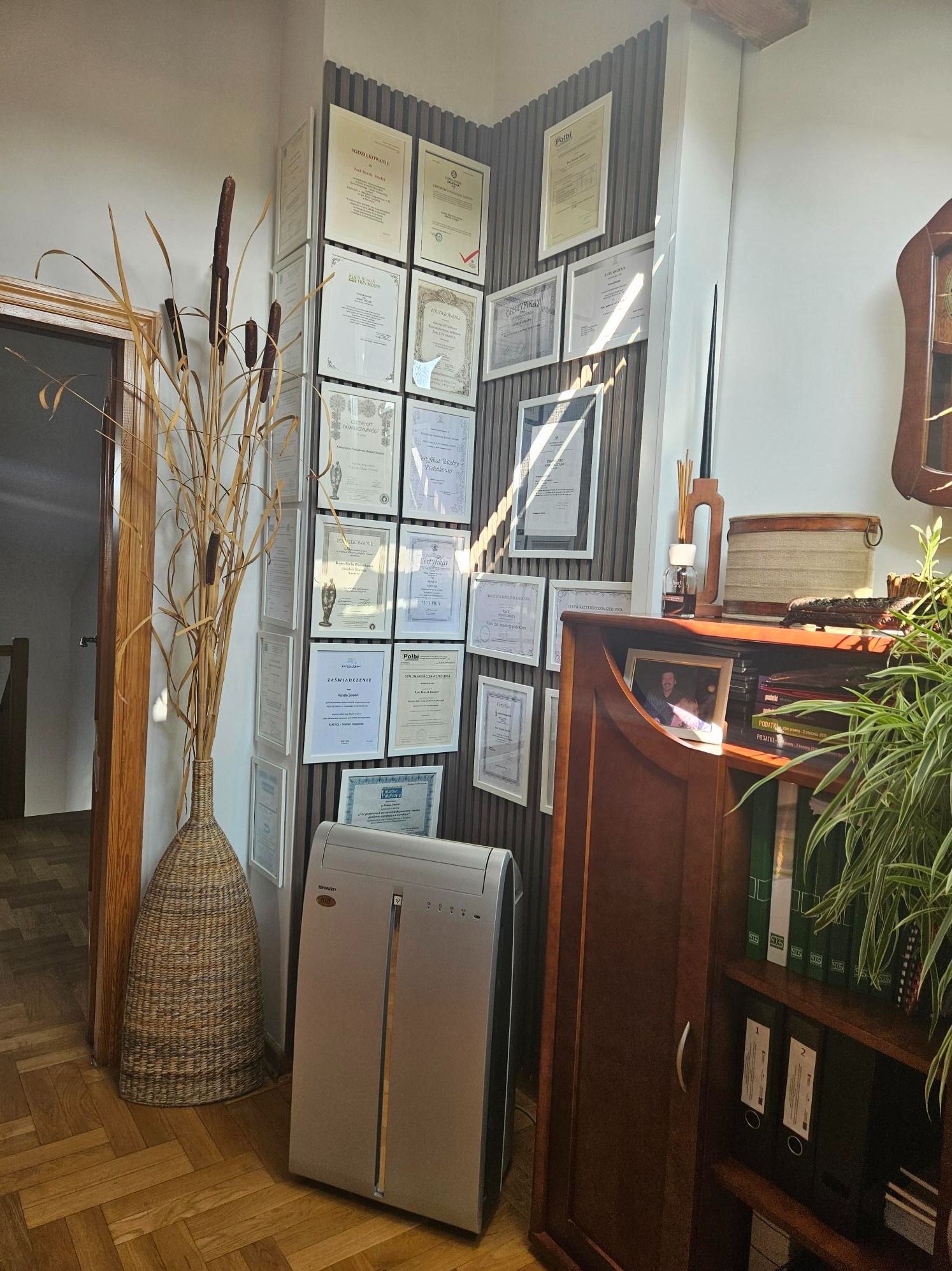 Ściana z certyfikatami i dyplomami w ramkach, obok regał z segregatorami i książkami o podatkach, trzcina w wazonie i niszczarka dokumentów w biurze