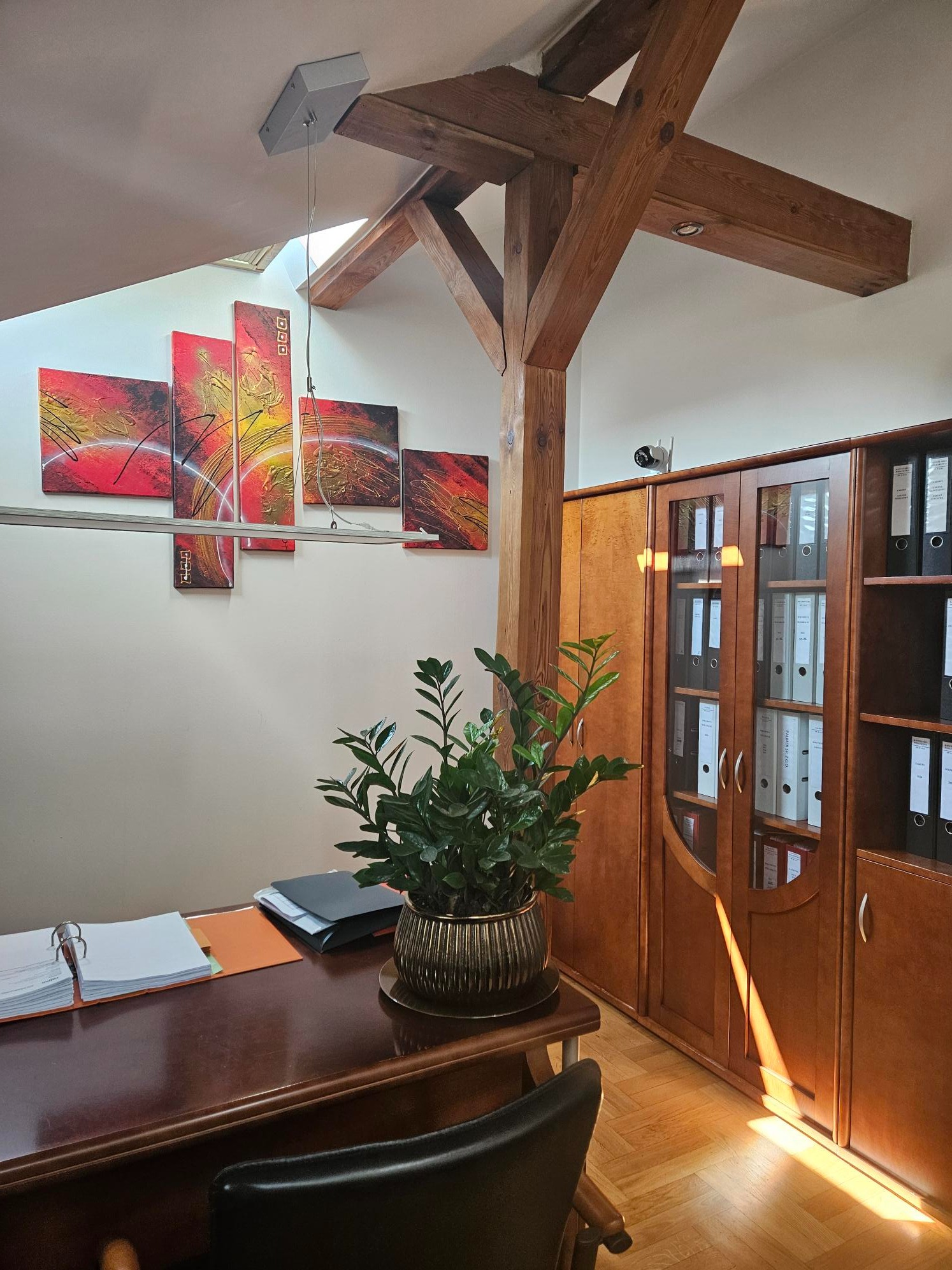 Biuro z drewnianą konstrukcją stropu, obrazami abstrakcyjnymi, szafą z segregatorami i kwiatem w ozdobnej doniczce na biurku