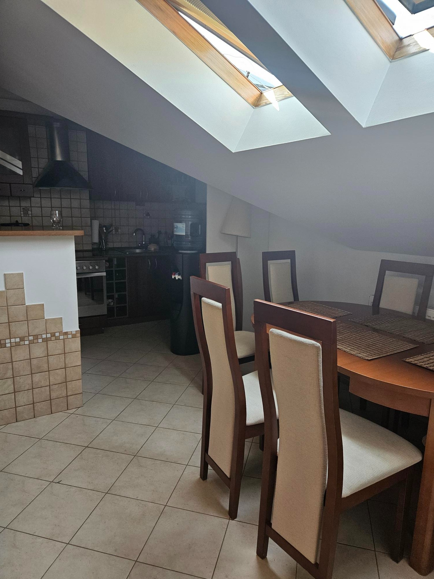 Umeblowana jadalnia z drewnianym stołem, krzesłami z beżową tapicerką i widokiem na aneks kuchenny pod skośnym dachem z oknami dachowymi.