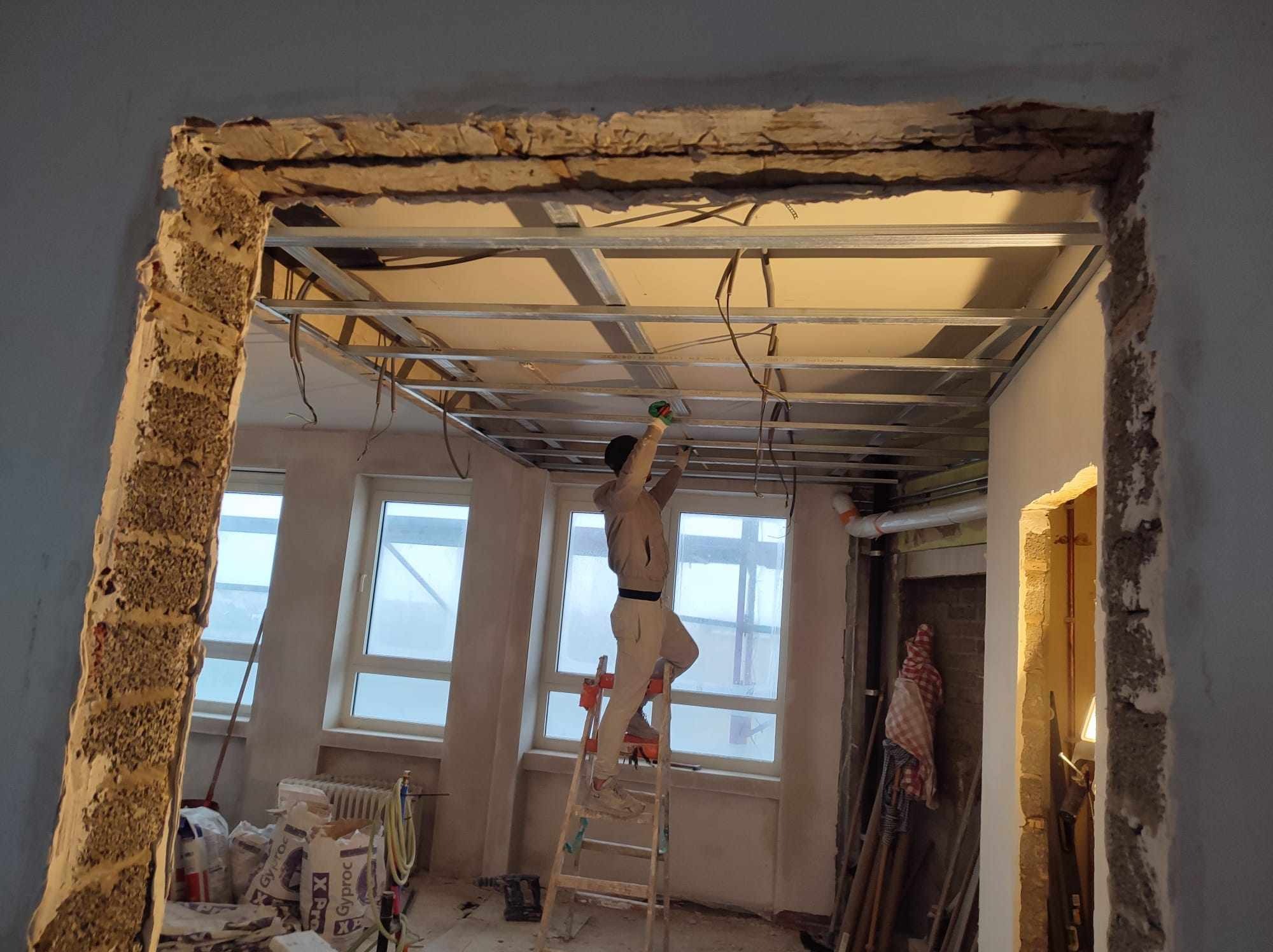 Pracownik na drabinie montuje metalową konstrukcję sufitu podwieszanego w remontowanym pomieszczeniu, widoczne worki z gipsem i surowy stan ścian.