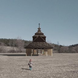 NASZA Pracownia Projektowa Piotr Krynicki - Wizualizacja drewnianej kapliczki z dwuspadowym dachem krytym gontem i krzyżem na szczycie, na otwartym polu, z sylwetką dziecka w oddali.