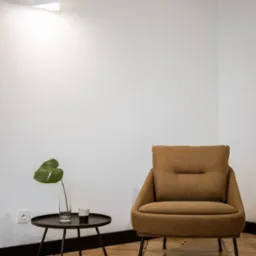 Beżowy fotel z poduszką, stolik kawowy z pojedynczym liściem monstery w wazonie i filiżanką, drewniana podłoga z geometrycznym wzorem, minimalistyczne oświetlenie na białej ścianie.