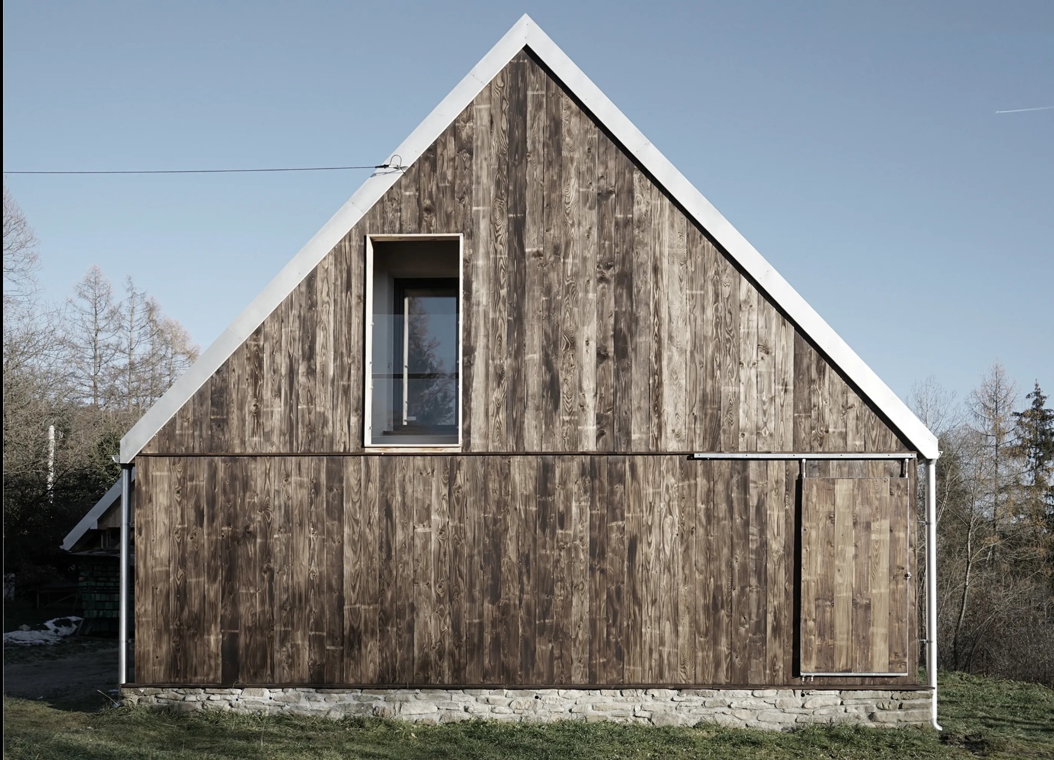 Nowoczesny dom o prostej bryle z drewnianą elewacją, akcentowany oknem z balustradą i przesuwanymi drzwiami, osadzony na kamiennej podmurówce.