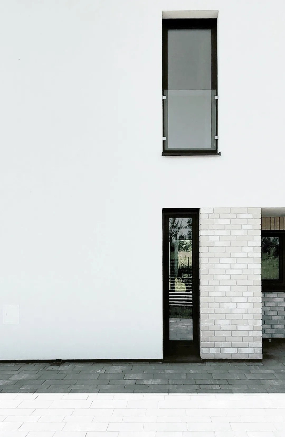 Minimalistyczna fasada budynku z białym tynkiem, wysokim oknem z barierką, przeszklonymi drzwiami wejściowymi i fragmentem elewacji z jasnej cegły.