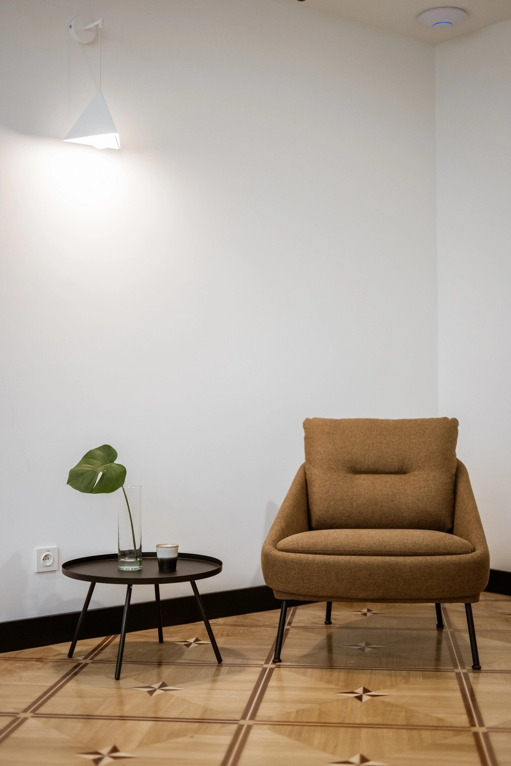 Beżowy fotel z poduszką, stolik kawowy z pojedynczym liściem monstery w wazonie i filiżanką, drewniana podłoga z geometrycznym wzorem, minimalistyczne oświetlenie na białej ścianie.