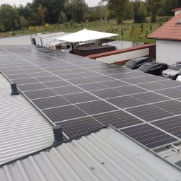 INSTAL-KON MACIEJ KONWA - Rozległa instalacja paneli słonecznych na blaszanym dachu, z pracownikiem w niebieskim kombinezonie leżącym obok, widoczne kominki wentylacyjne i zaparkowane samochody w tle.