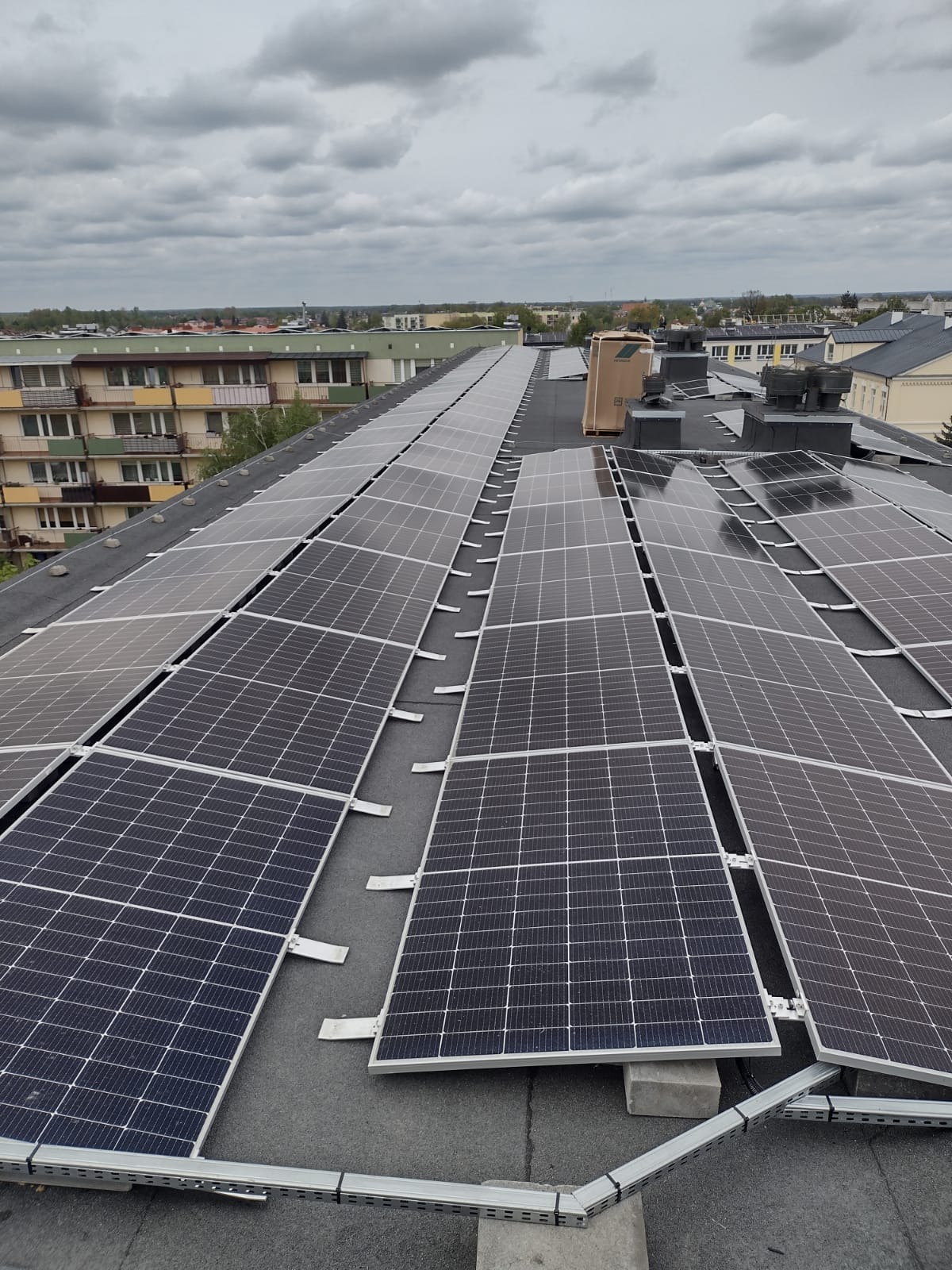 Rząd paneli słonecznych na płaskim dachu budynku z widocznymi elementami montażowymi i kartonowym pudełkiem w tle, pochmurne niebo.