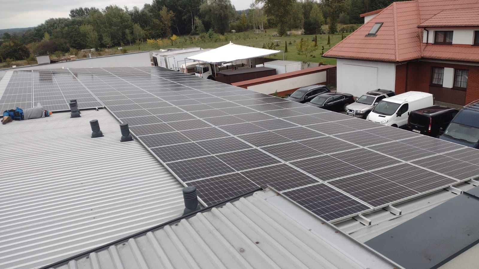 Rozległa instalacja paneli słonecznych na blaszanym dachu, z pracownikiem w niebieskim kombinezonie leżącym obok, widoczne kominki wentylacyjne i zaparkowane samochody w tle.