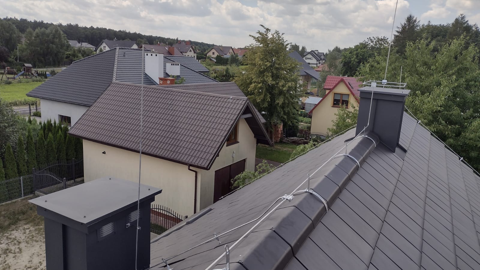 Antena odgromowa zamontowana na kominie i dachu z dachówki ceramicznej na tle zabudowy jednorodzinnej w słoneczny dzień.