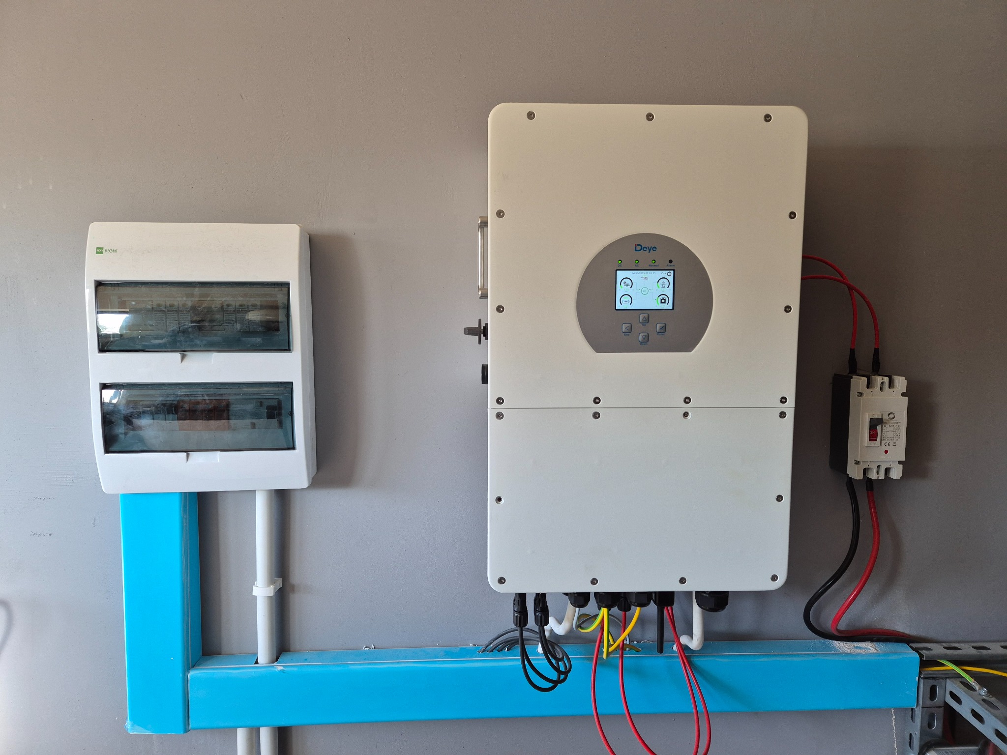 Biały inwerter sieciowy Deye z wyświetlaczem i podłączonymi kablami, obok skrzynka bezpiecznikowa z widocznymi zabezpieczeniami, całość zamontowana na szarej ścianie, z niebieską listwą przypodłogową.