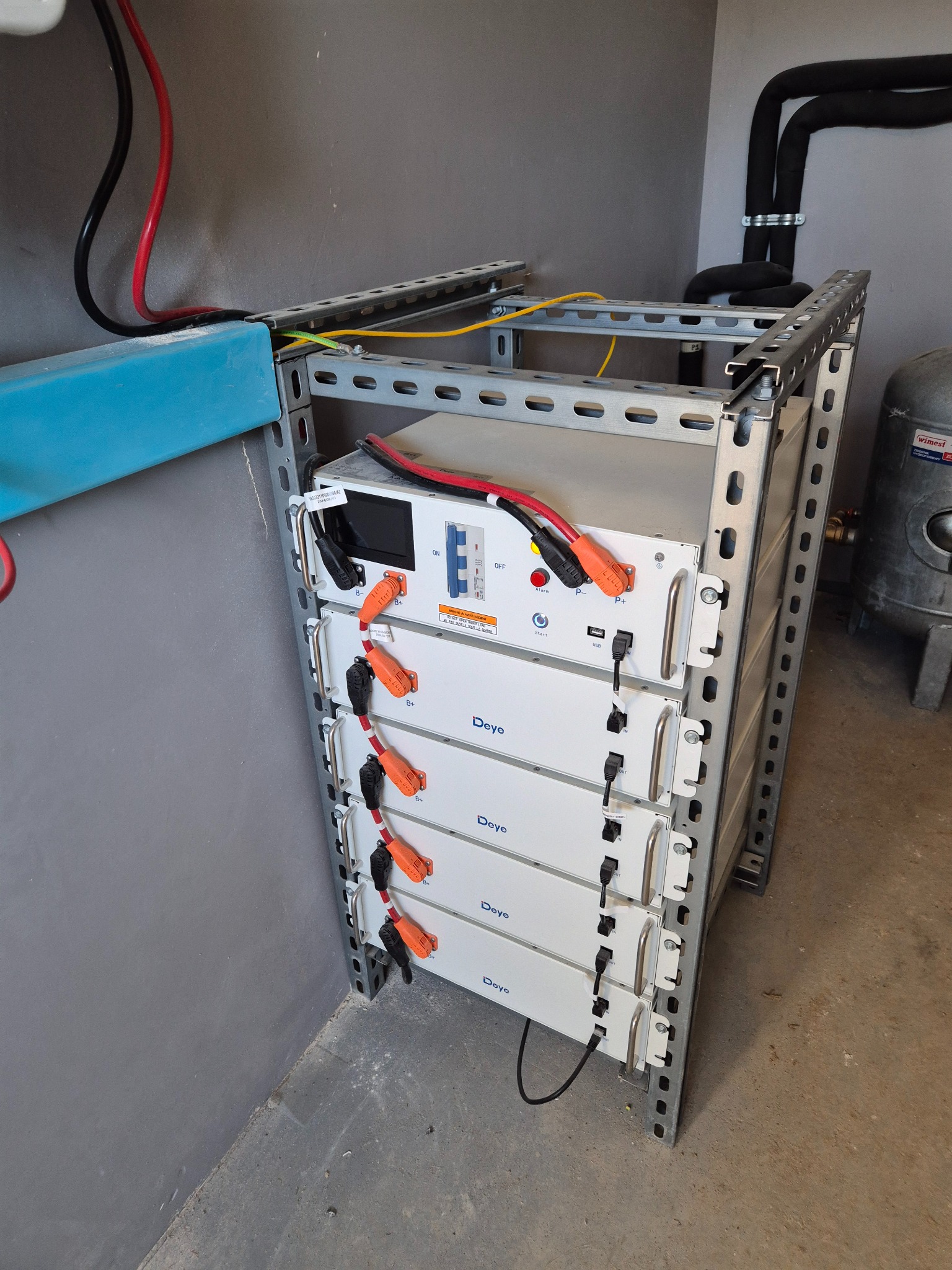 Bateria magazynu energii marki Deye zamontowana na metalowym regale w pomieszczeniu technicznym z rurami i zbiornikiem wyrównawczym w tle.
