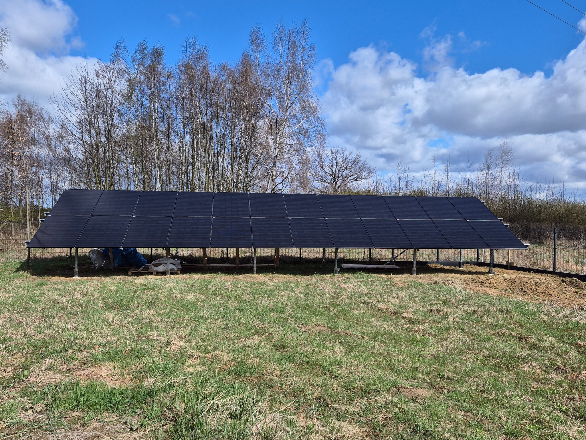 Panel słoneczny na metalowej konstrukcji w plenerze, widoczne worki i plandeka pod panelami, w tle drzewa i błękitne niebo z chmurami.