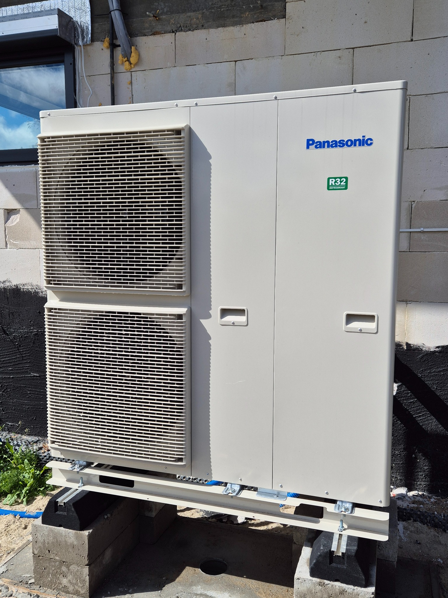 Zewnętrzna jednostka pompy ciepła Panasonic R32 zamontowana na betonowych blokach, widoczna instalacja i przewody na tle ściany z betonu komórkowego.
