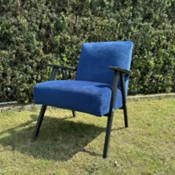 Niebieski fotel tapicerowany z czarnymi drewnianymi nogami stoi na trawie przed zielonym żywopłotem. Fotel w stylu retro, odnowiony.