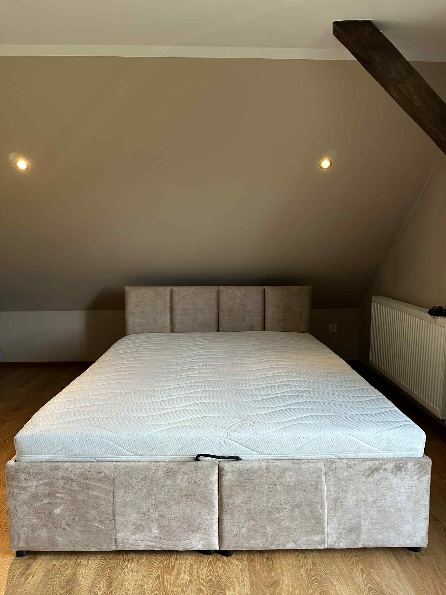 Beżowe łóżko tapicerowane z pojemnikiem na pościel i materacem w pokoju na poddaszu, widoczne drewniane belki stropowe i grzejnik.